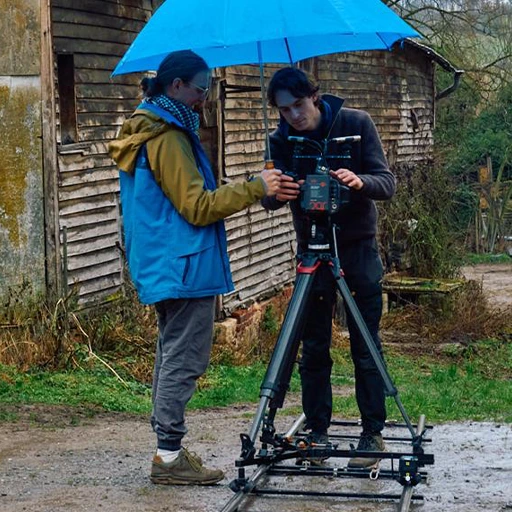 A photo of Rick and Daan with a Blackmagic URSA Cine Immersive camera on a dolly on a set of tracks on an unpaved outdoor street. A large umbrella is next to them.